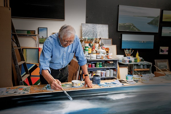 A man standing over a tabled canvas and paint points, putting down brush strokes with a paintbrush